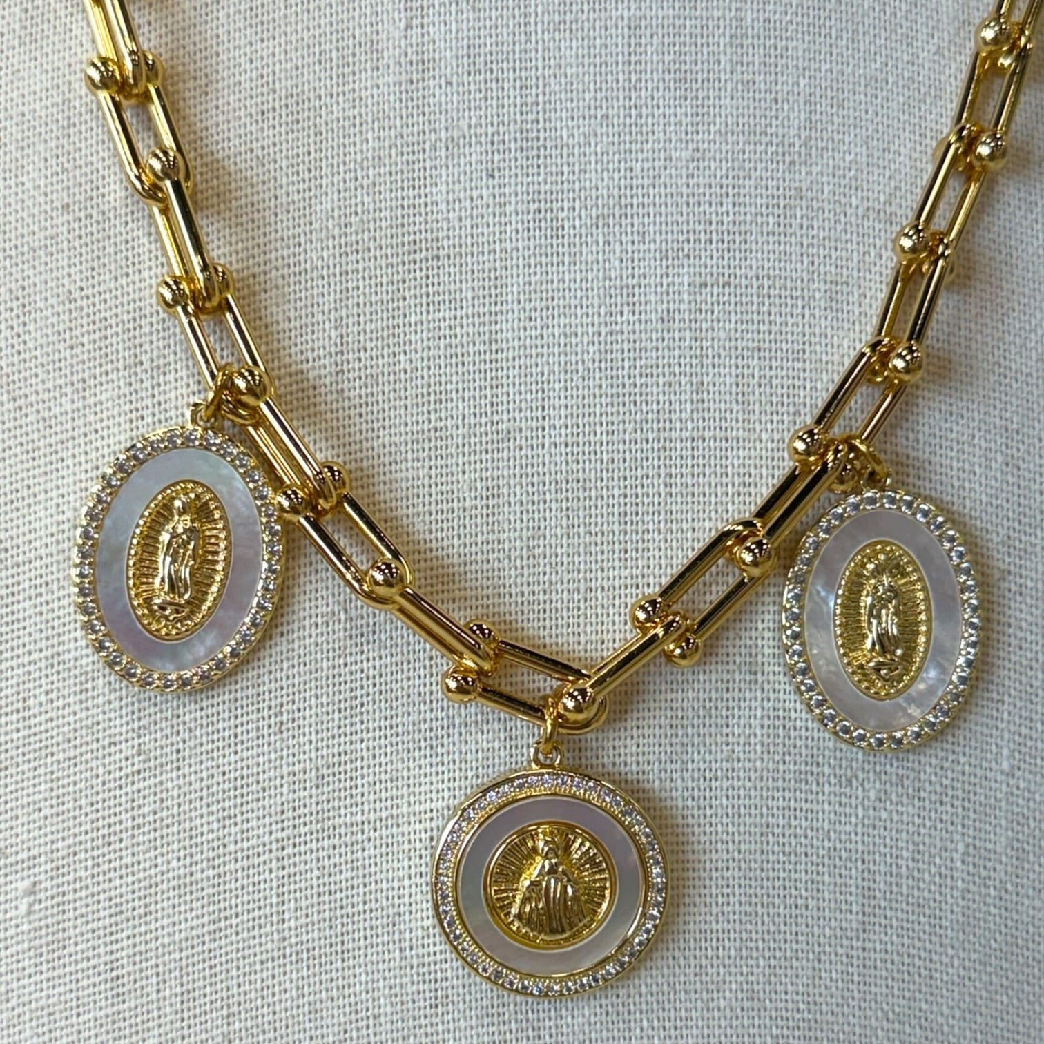 Gold chain necklace with three oval pendants featuring gold Miraculous Medal motifs set in white shell-like centers and bordered with small clear stones, displayed on a textured off-white fabric background.
