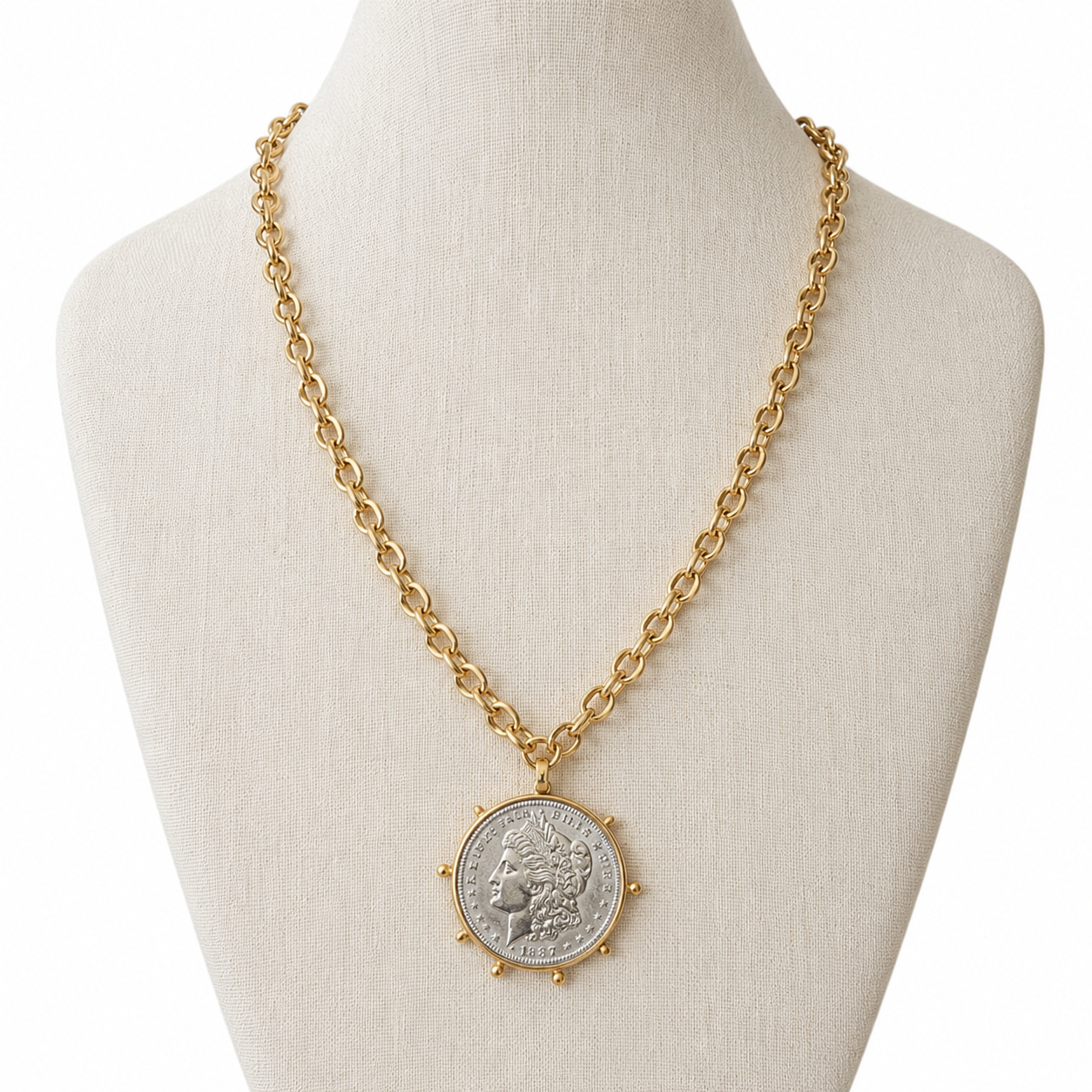 A gold chain necklace with a circular silver coin pendant, displayed on a cream fabric jewelry bust against a white background.