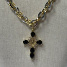 A gold and silver chain necklace with a cross pendant featuring black teardrop-shaped stones and a central gold-toned oval detail, displayed on a textured beige fabric background.