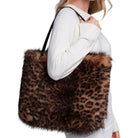 A woman wearing a white sweater carries a large faux-fur tote bag with a brown and black leopard print pattern against a plain white background.