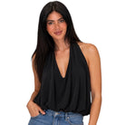 A woman with long dark hair wearing a black halter-neck top with a deep V-neckline and draped fabric, paired with blue jeans, against a solid white background.