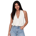 A woman modeling a sleeveless cream halter-style top with a deep V-neck and draped neckline, paired with light-wash blue jeans, against a plain white background.