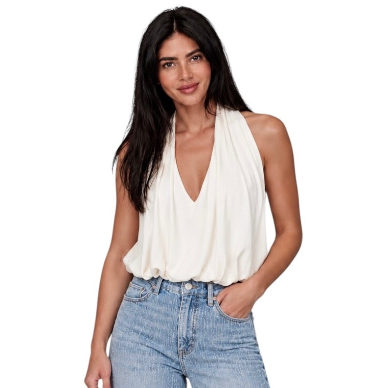 A woman modeling a sleeveless cream halter-style top with a deep V-neck and draped neckline, paired with light-wash blue jeans, against a plain white background.