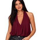 A woman wearing a sleeveless halter-style top in deep burgundy with a draped V-neckline, paired with black leather pants, against a plain white background.