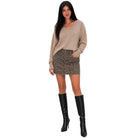 A woman modeling a beige long-sleeve knit sweater, a brown and black leopard-print mini skirt, and black knee-high heeled boots, set against a plain white background.