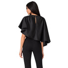 A woman shown from the back wearing a black draped cape-style top with layered ruffle edges and a small keyhole cutout at the neckline, paired with black leggings, against a plain white background.