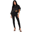 A woman modeling a black draped cape-style top and black leggings with black high-heeled sandals against a plain white background.