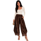 A woman modeling high-waisted, wide-leg brown velvet pants with lace trim at the hems, paired with a cream long-sleeve top and holding a small woven clutch, against a solid white background.