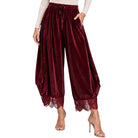 A model wears high-waisted, balloon-style velvet harem pants in deep burgundy with a lace hem and a drawstring waist, paired with a cream long-sleeve top and metallic high-heeled sandals against a plain white background.
