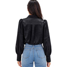 A woman viewed from the back wearing a black long-sleeve blouse with textured fabric and puffed shoulders, paired with light blue jeans, against a plain white background.