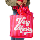 A person holding two Christmas-themed shopping bags against a white background. The larger bag in front is pink and red vertically striped with the words 'Very Merry' in white script, and the smaller bag behind it is red with pink handles and a pink bow.