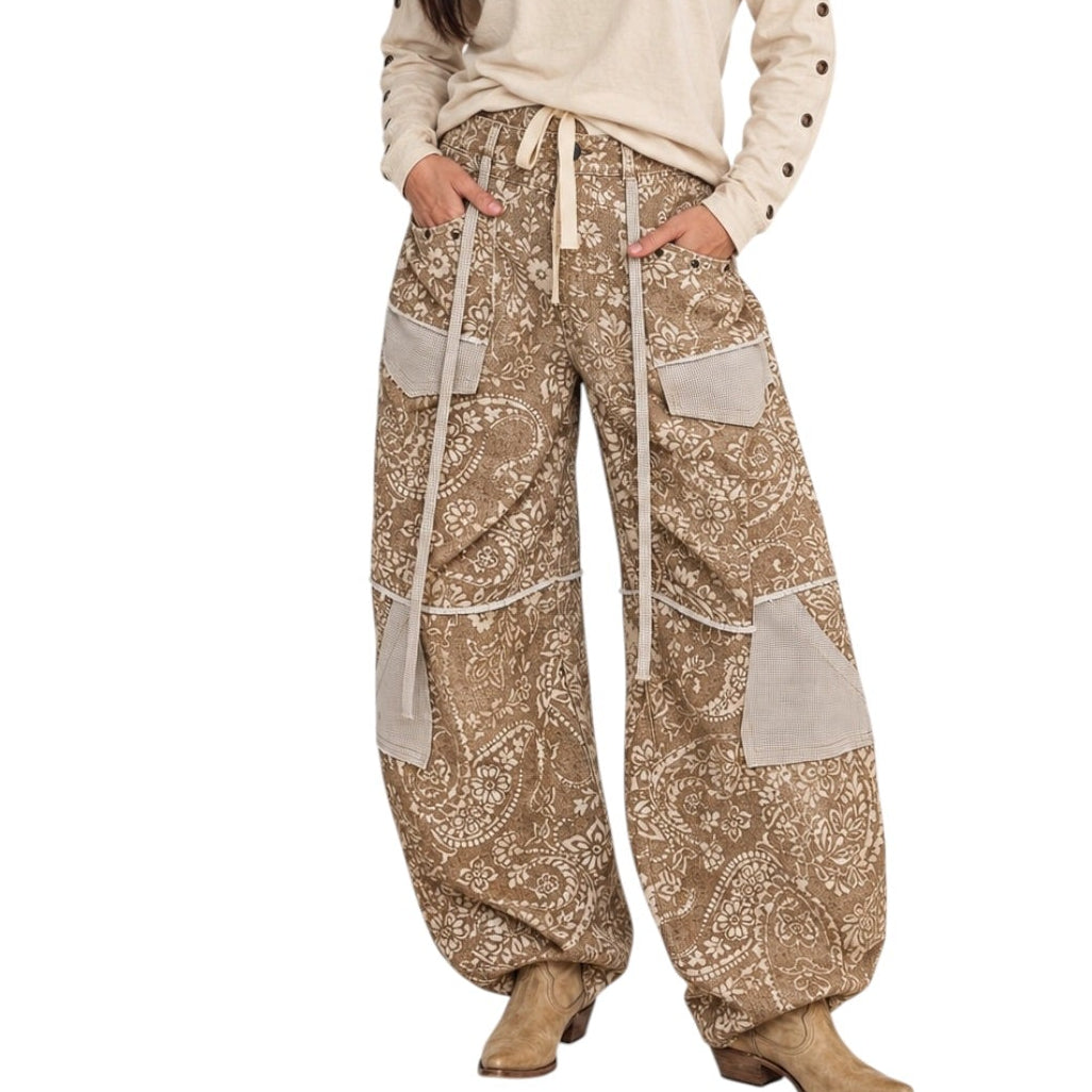 A woman modeling a paisley-style outfit featuring a cream off-the-shoulder long-sleeve top with metal studs and wide-leg beige paisley pants with contrast panels and a drawstring waist, paired with tan boots, against a plain white background.