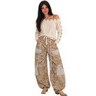 A woman modeling a bohemian-style outfit featuring a cream off-the-shoulder long-sleeve top with metal studs and wide-leg beige paisley pants with contrast panels and a drawstring waist, paired with tan boots, against a plain white background.