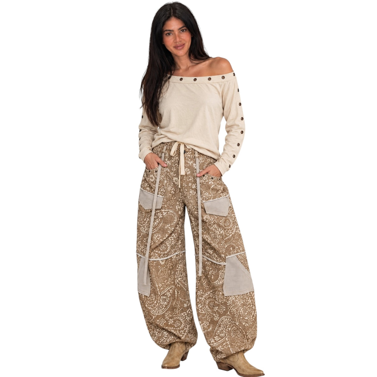 A woman modeling a bohemian-style outfit featuring a cream off-the-shoulder long-sleeve top with metal studs and wide-leg beige paisley pants with contrast panels and a drawstring waist, paired with tan boots, against a plain white background.