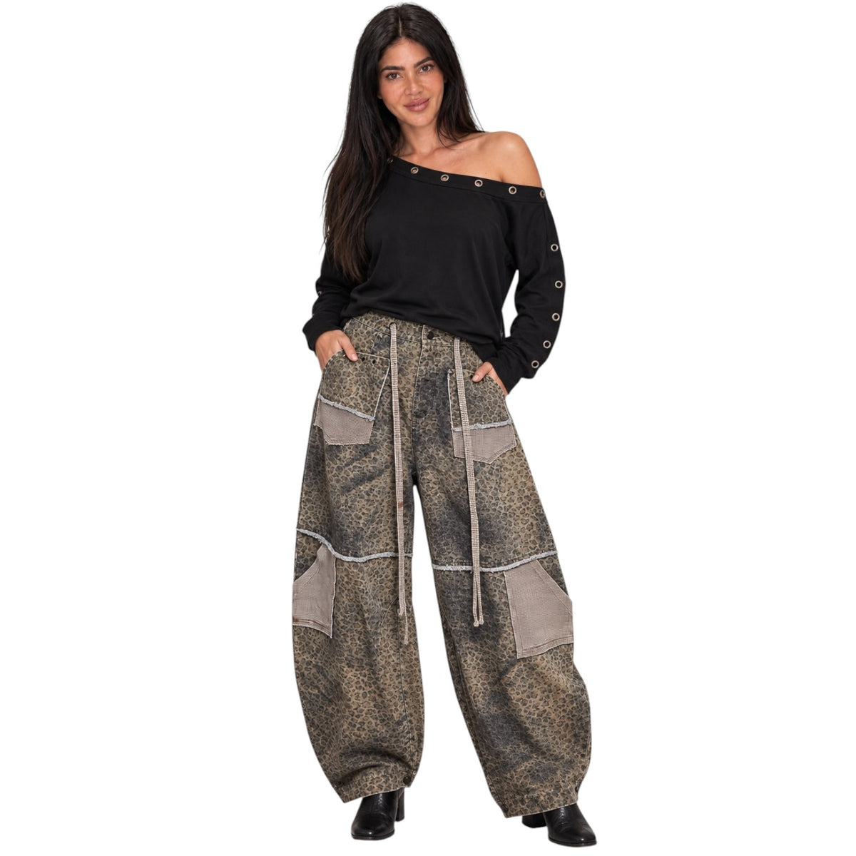 A woman modeling a pair of loose, wide-leg patchwork pants in a brown and beige animal print with a beige drawstring, paired with a black off-the-shoulder long-sleeve top and black shoes, against a plain white background.