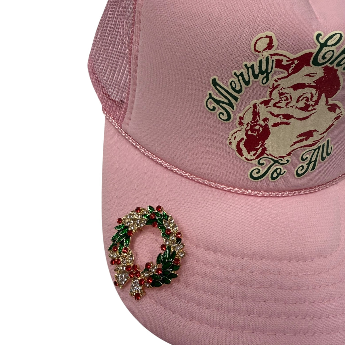 A light pink trucker hat featuring a red and green embroidered Santa Claus graphic on the front and a red and green rhinestone Christmas wreath embellishment on the brim.