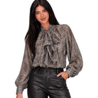 A woman modeling a sheer long-sleeve blouse with a brown and beige animal print and a large ruffled bow at the neck, paired with black leather-look pants against a plain white background.