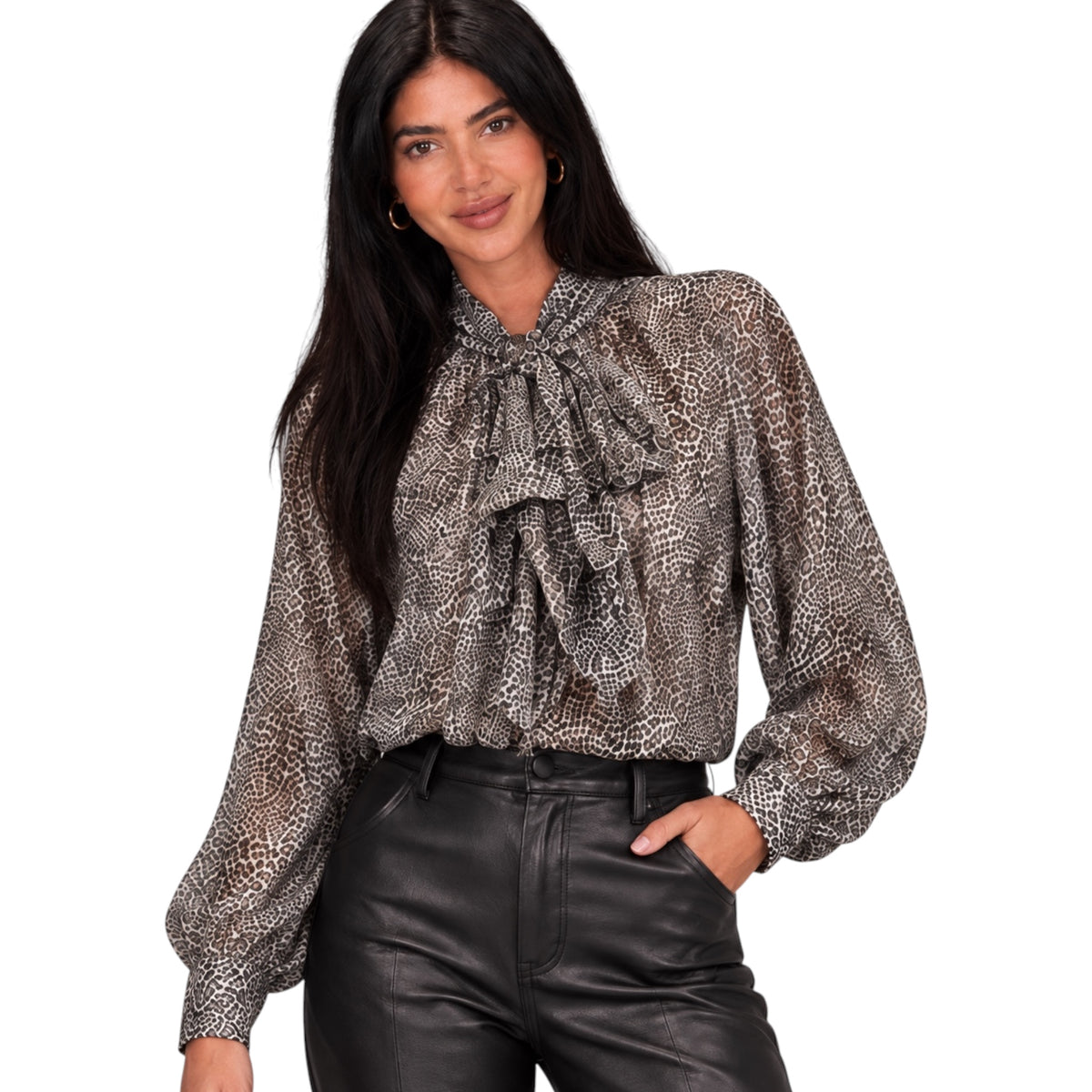 A woman modeling a sheer long-sleeve blouse with a brown and beige animal print and a large ruffled bow at the neck, paired with black leather-look pants against a plain white background.