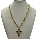 A gold-tone chain necklace with a cross pendant featuring black stones, displayed on a beige fabric jewelry bust against a white background.