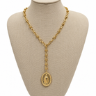 A gold-toned lariat-style necklace with a large oval pendant featuring a religious figure, displayed on a beige fabric jewelry bust against a white background.