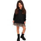 A young girl modeling a black long-sleeve hoodie dress with a leopard-print ruffled skirt and black combat boots, set against a plain white background.
