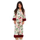 A young girl modeling a two-piece holiday pajama set with a festive print of red bows, green leaves, and red deer on a cream background, featuring dark red faux-fur cuffs and hem on the pants.