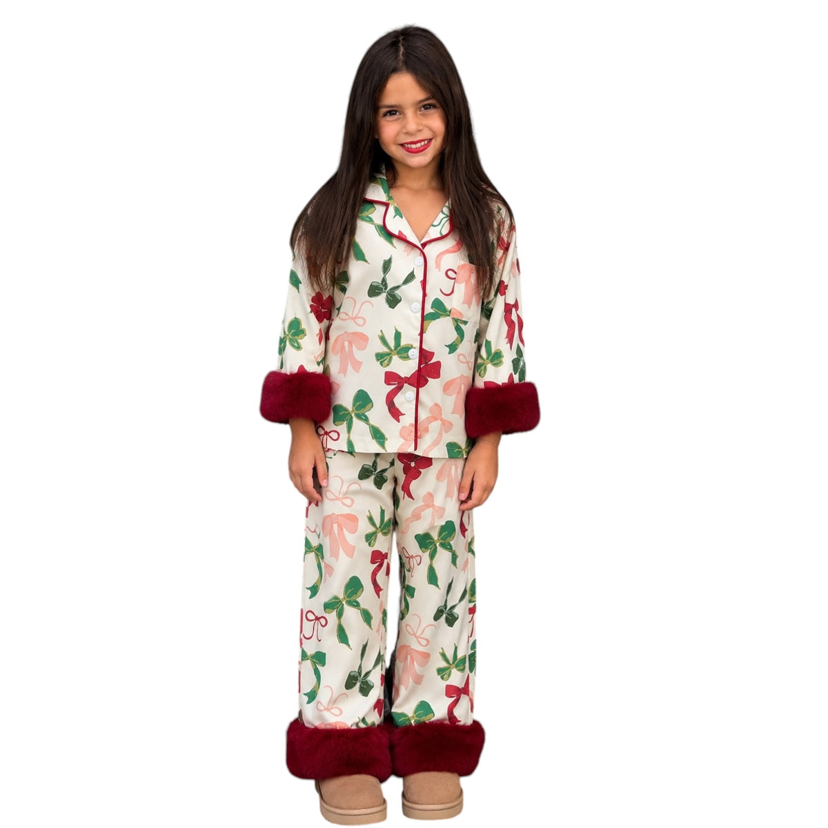 A young girl modeling a two-piece holiday pajama set with a festive print of red bows, green leaves, and red deer on a cream background, featuring dark red faux-fur cuffs and hem on the pants.