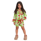 A young girl modeling a light green Grinch-themed pajama set with red bows and green-and-yellow Grinch prints, paired with tan slip-on shoes, on a solid white background.