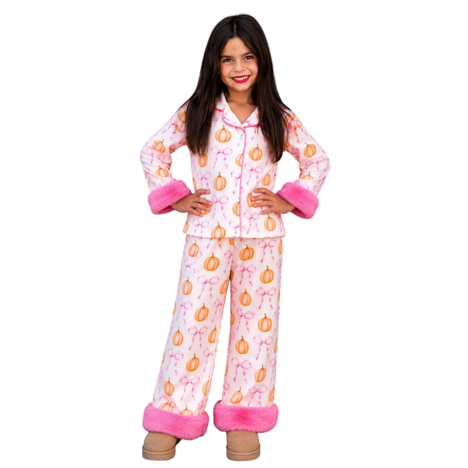 A young girl modeling a two-piece pajama set featuring an orange pumpkin and pink bow pattern on a white background, with pink faux-fur cuffs and hem, set against a plain white studio background.