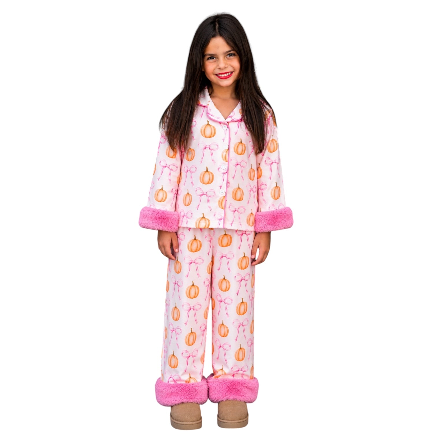 A young girl stands against a plain white background wearing a two-piece pajama set. The pajamas are white with an all-over orange pumpkin print and pink bows, finished with bright pink faux-fur trim at the cuffs and ankles.