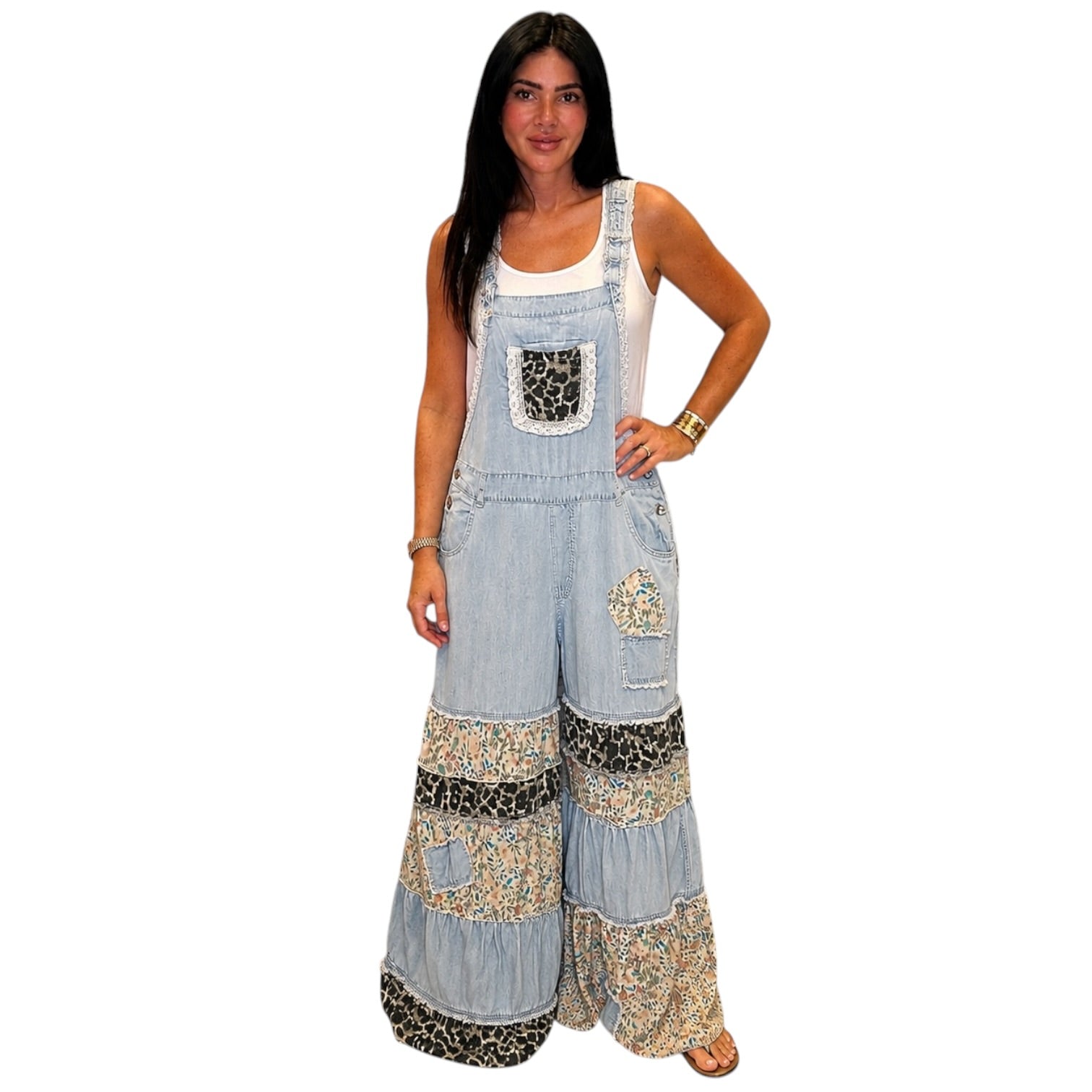 A woman modeling a light blue denim overall-style jumpsuit with wide legs and mixed patterned patchwork details, worn over a white tank top against a plain white background.