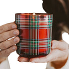 A close-up of a hand holding a red glass candle with a green, white, and blue plaid pattern. The brand name 'SWEET TREATS & DECOR' is embossed at the bottom of the glass.