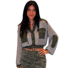 A woman modeling a cropped button-front knit top with striped gray and olive green panels, green patch pockets, and green elbow patches, paired with camouflage pants, against a plain white background.