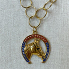 A gold-tone horse head pendant necklace set within a horseshoe shape, decorated with red and blue enamel and small rhinestones, shown on a textured white fabric background.