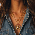 A close-up of a model wearing a gold-tone chain necklace with a horse head pendant and a red-and-gold horseshoe design, styled with a blue denim shirt.