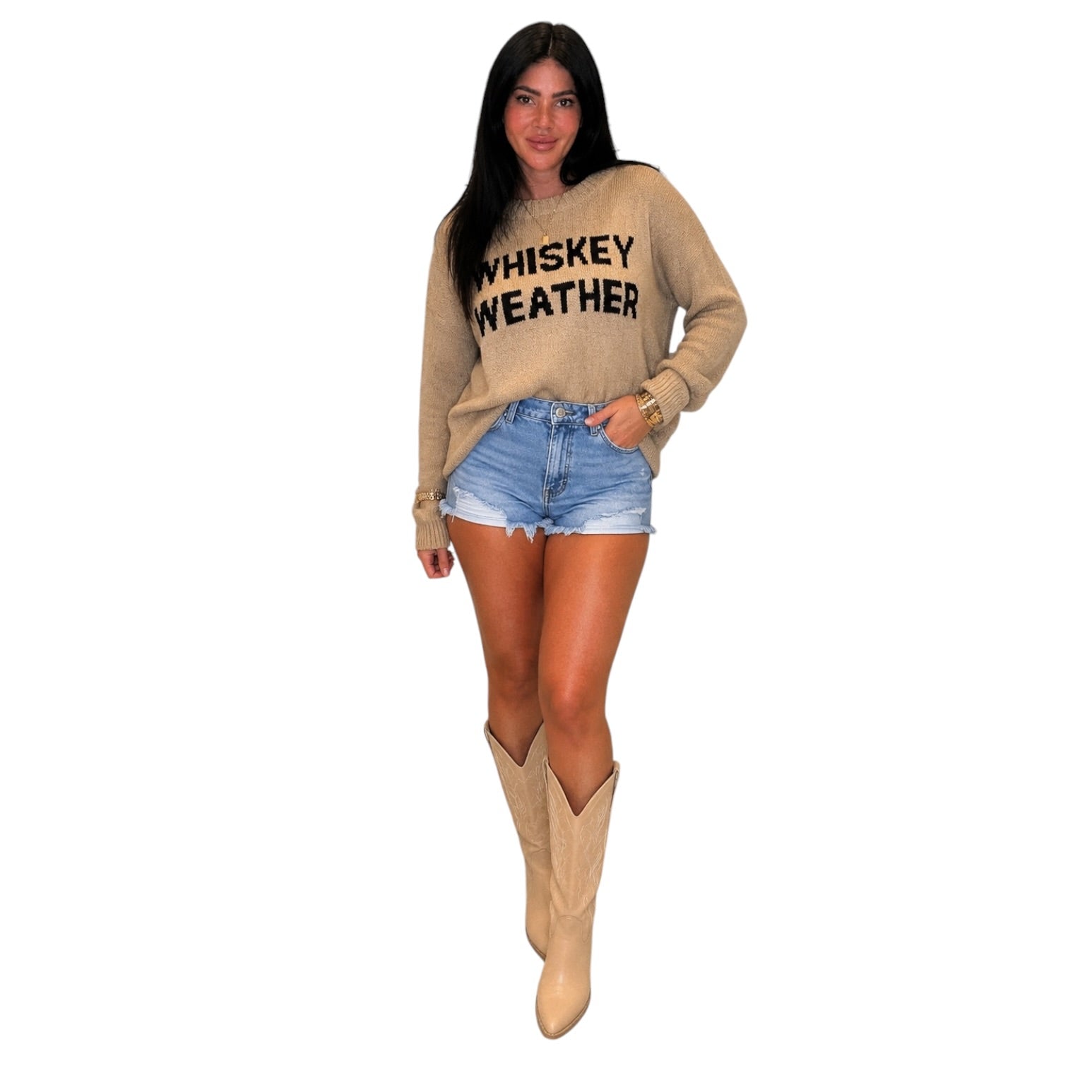 A woman modeling a beige crewneck sweater with 'WHISKEY WEATHER' printed in black across the chest, paired with denim shorts and beige cowboy boots against a plain white background.