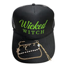 A black trucker hat with 'Wicked Witch' printed in neon green on the front, styled with a gold chain brooch featuring star and moon charms and a pearl bead chain on the brim.