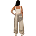 A woman modeling a pair of beige and brown animal print patchwork harem pants with a drawstring waist, paired with a cream crop top and white sneakers, against a plain white background.