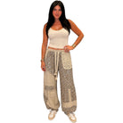 A woman modeling a pair of beige and brown animal print patchwork harem pants with a drawstring waist, paired with a cream crop top and white sneakers, against a plain white background.