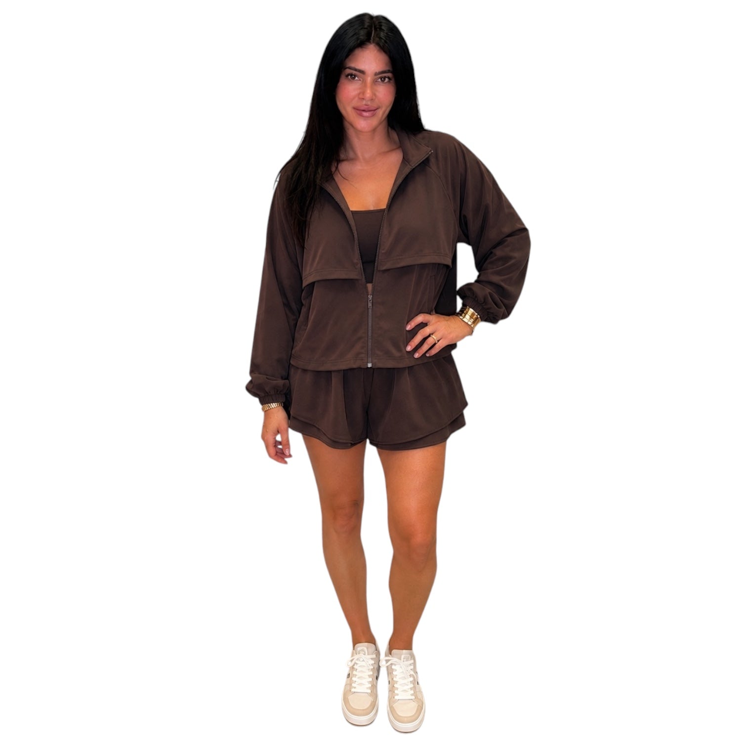 A woman modeling a dark brown two-piece matching set consisting of a lightweight zip-up jacket and matching shorts, paired with white sneakers, against a plain white background.