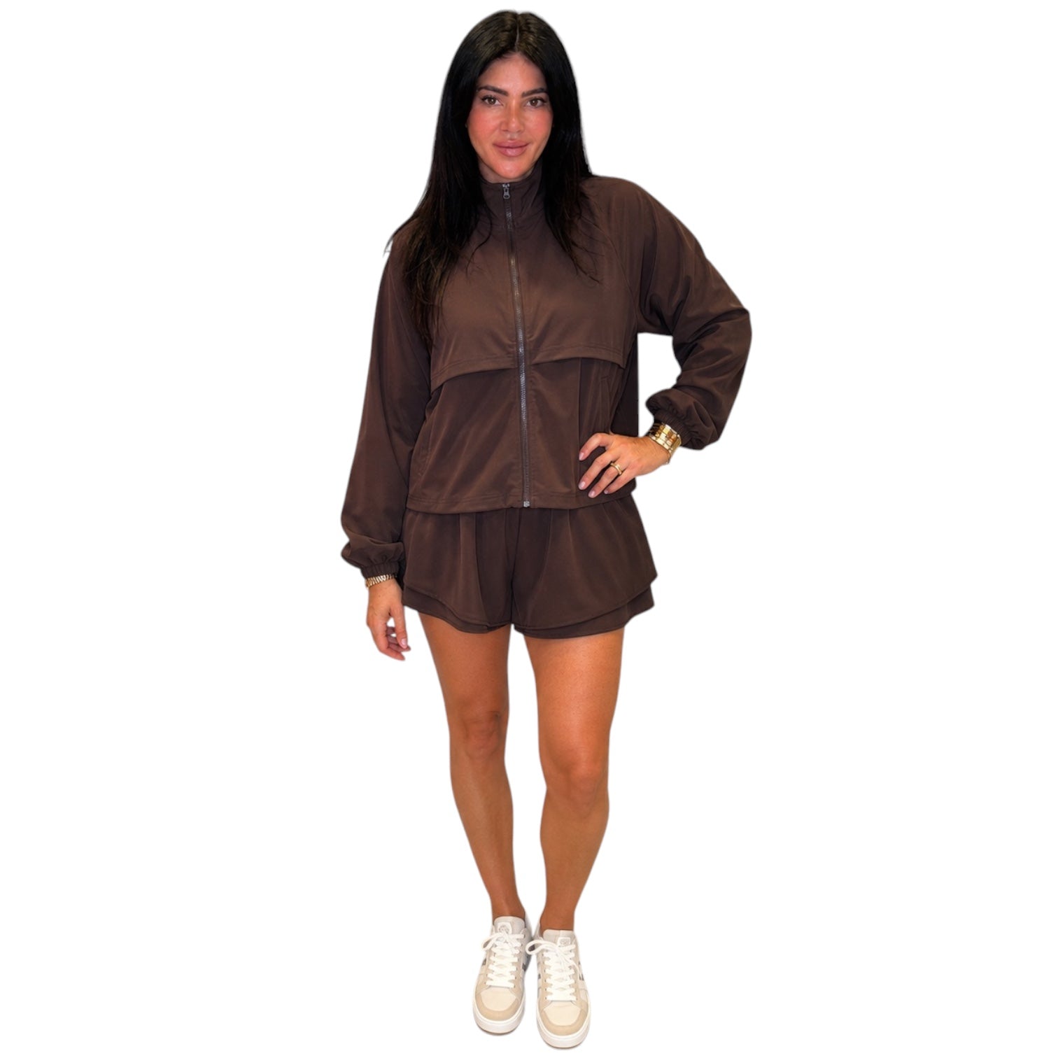 A woman modeling a matching brown zip-up jacket and shorts set, paired with white sneakers, against a plain white background.