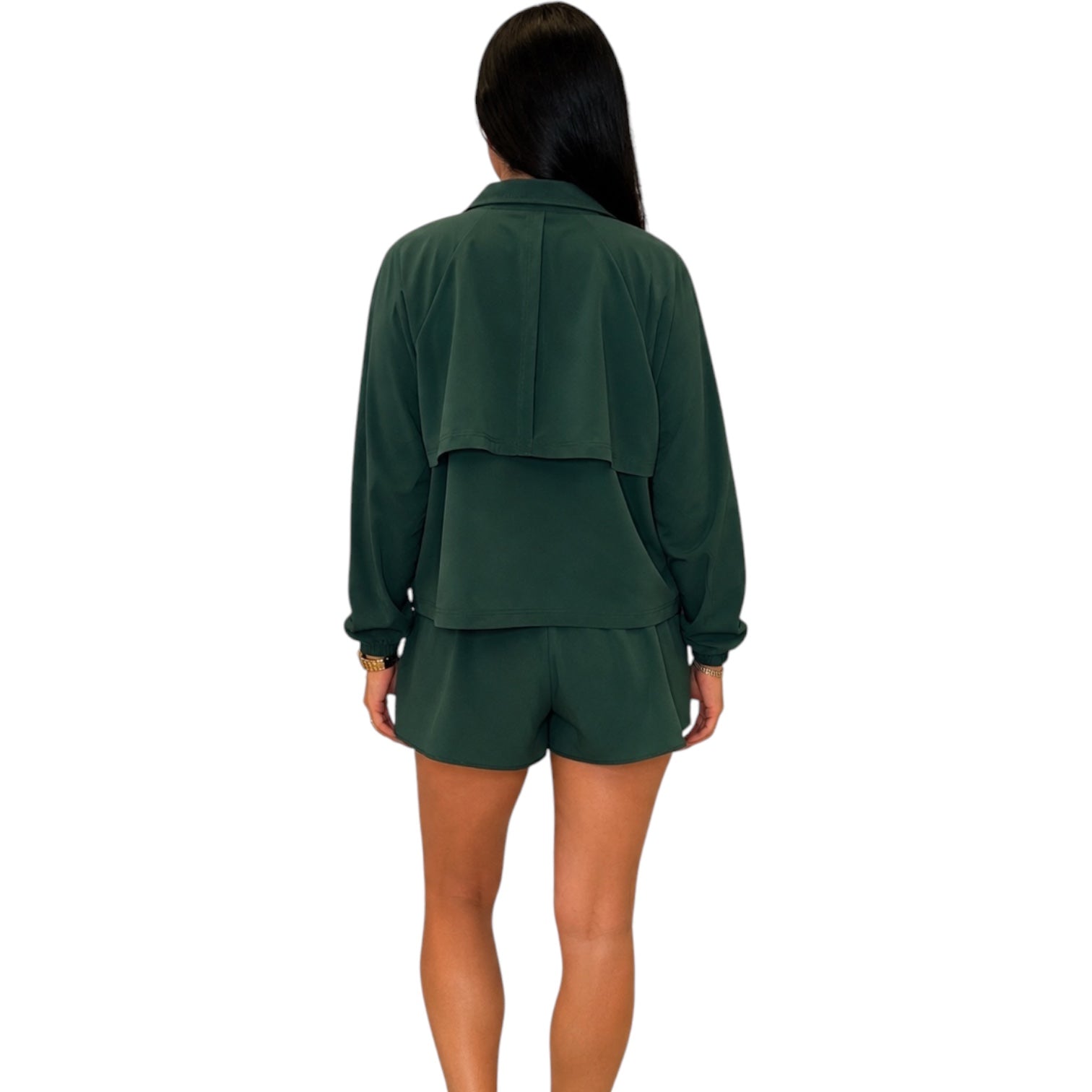 A woman viewed from the back wearing a dark green long-sleeve cropped jacket with a layered back detail and matching dark green shorts, set against a plain white background.