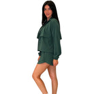 A woman modeling a dark green two-piece lounge set consisting of a long-sleeve cropped top with layered ruffle details and matching shorts, set against a plain white background.