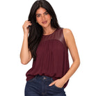 A woman modeling a sleeveless top with a deep burgundy body and a darker burgundy faux-leather yoke across the shoulders, paired with dark blue jeans against a plain white background.