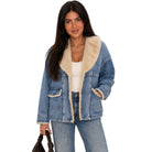 A woman modeling a light blue denim jacket with a thick cream-colored shearling lining on the collar, cuffs, and pockets, paired with blue jeans and a white top against a plain white background.