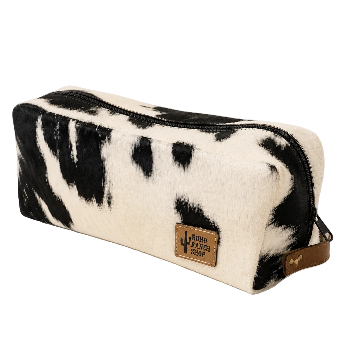 A rectangular zippered pouch with a black and white cowhide pattern, featuring a brown faux-leather brand patch that reads 'NORTH RANCH SHOP' with a cactus logo, set against a clean white background with dried grass in the corner.
