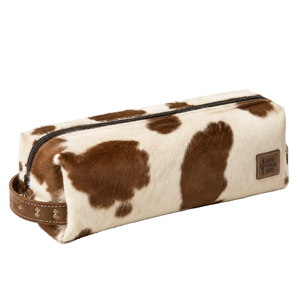 A cowhide zippered pouch with brown and cream spots, a brown leather strap, and a small brown brand patch on the side, photographed on a clean white background with dried grass decor in the corner.