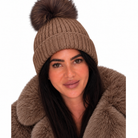 A woman wearing a brown ribbed knit beanie with a large matching faux-fur pom-pom and a brown fur-trim coat, shown against a plain white background.