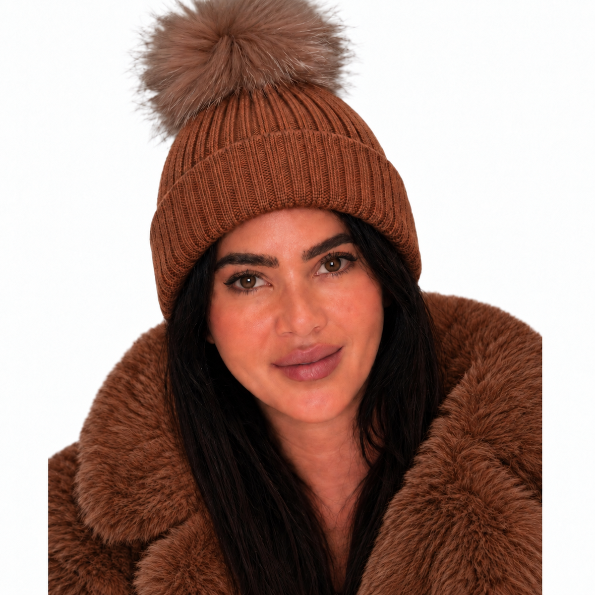 A woman wearing a ribbed brown knit beanie with a large matching faux-fur pom-pom, paired with a brown faux-fur collar jacket, against a plain white background.