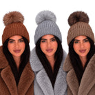 Three women wearing ribbed knit pom-pom beanies in brown, light gray, and taupe, paired with faux fur coats against a plain white background.